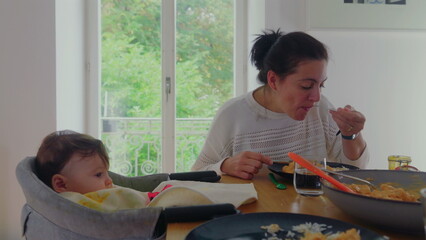 Mother carefully feeding baby in high chair, baby eating food with mother’s assistance, intimate...