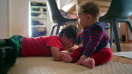 Baby sitting on the floor with older sibling nearby, baby smiling with excitement, sibling...