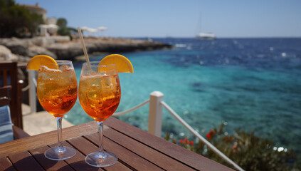 Two aperol spritz cocktails with orange slices on wooden table, turquoise sea and rocky coast in background, bright summer day with clear sky, wide angle vacation scene