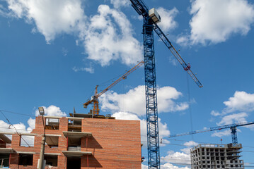 A large number of tower construction cranes are involved in the construction of a new residential area. Construction equipment. Tower cranes among the trees and the blue sky with clouds.