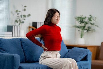Asian woman having backache, touching her back with hand, suffering from back pain, sitting on blue sofa in living room
