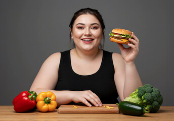 Woman holding hamburger at table with fresh vegetables – concept of balanced lifestyle, food choices, or contrast between healthy eating and indulgence.