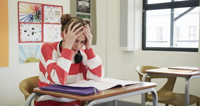In classroom, girl with headphones looking stressed over open notebook, at school