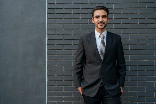 Confident young businessman standing in a formal black suit and tie against a dark brick wall, posing with a professional and self assured expression. - Powered by Adobe