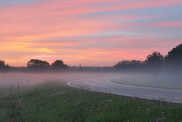 A serene rural road at sunrise with a misty foreground, silhouetted trees, and a pink-orange sky full of scattered clouds.