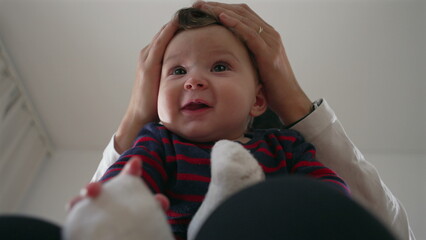 Mother holding baby’s head gently, playful interaction as baby smiles while sitting on her lap,...