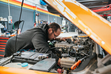 Focused male mechanic leaning into the open hood of an orange vehicle during intensive engine inspection and repair in an automotive shop
