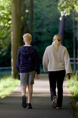 A boy and a girl are walking down a walkway in a forest with the sun shining in from the side.