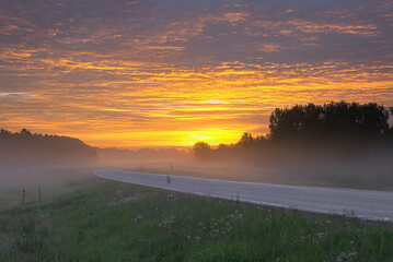 A glowing golden sunrise lights up a foggy rural road with trees silhouetted against the mist and a vivid yellow-orange sky.