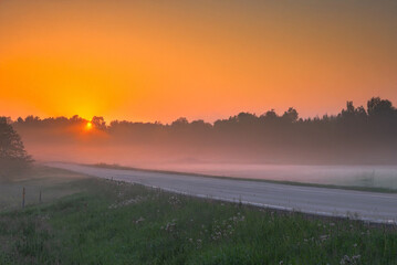 A glowing golden sunrise lights up a foggy rural road with trees silhouetted against the mist and a vivid yellow-orange sky.