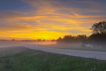 A glowing golden sunrise lights up a foggy rural road with trees silhouetted against the mist and a vivid yellow-orange sky.