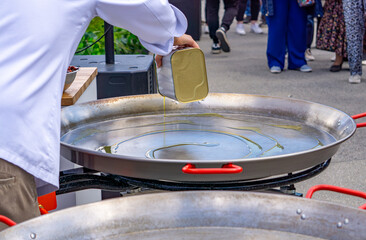 chef pours oil onto a large metal pan in preparation for outdoor cooking. The scene is part of a...