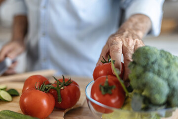 Senior man in kitchen cooking healthy meal. Handsome senior man is cooking in the kitchen and smiling while standing at table full with fresh colorful vegetables.