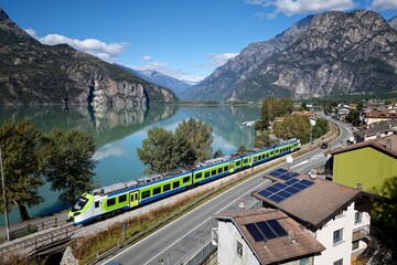 A vibrant train glides beside Lago di Mezzola, mirroring the calm lake and towering Alps, as sunlight dances on rooftops and crystal-clear waters in northern Italy&rsquo;s tranquil paradise.