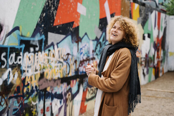 Cheerful woman with curly hair standing near colorful graffiti wall in the city