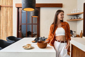 Young woman in an orange shirt and white pants, smiling while preparing healthy food in a stylish kitchen, emphasizing a cozy lifestyle and seasonal freshness