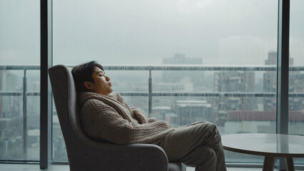 Solitude and Contemplation:  A person sits in a chair by a window, enveloped in thoughtful contemplation, as they gaze upon a cityscape during rainy weather. 