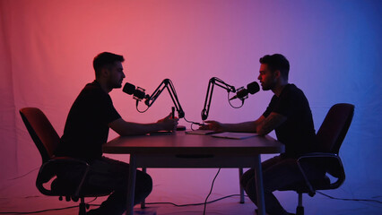 Podcast Recording Session: Two individuals engaged in a captivating podcast, seated across a table in a studio with microphones poised, bathed in a spectrum of vibrant hues.