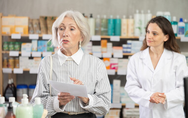 Mature woman stands in a European pharmacy with a medical prescription and consults a pharmacist about the choice of pills and medicines