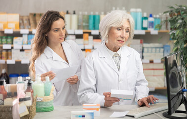 Obraz premium Senior female pharmacist helps a female assistant work with a computer in a pharmacy. They enter new arrivals of pills for head, migraine and allergy. Pharmacists work in a pharmacy