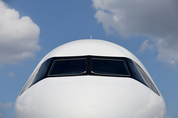 Front view of a commercial airplane cockpit with reflections on fuselage against blue ski and white clouds.