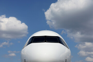 Front view of a commercial airplane cockpit with reflections on fuselage against blue ski and white clouds.
