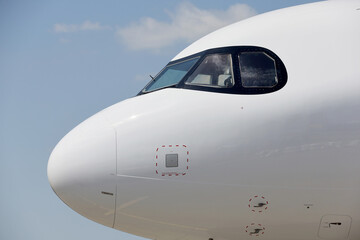 Lateral view of a commercial airplane cockpit with reflections on fuselage against blue ski and white clouds.