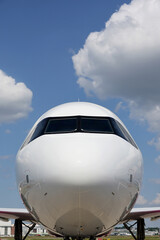 Front view of a commercial airplane cockpit with reflections on fuselage against blue ski and white clouds.