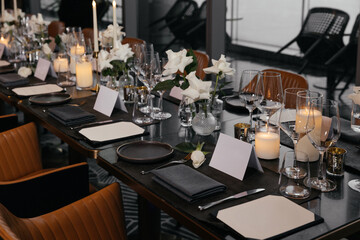  Panoramic view of a refined black table setting with white roses, handmade plates, natural wood chairs, and glasses. Elegant decor with fresh flowers for a sophisticated event atmosphere.