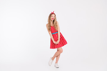 Happy young woman in a red polka-dot dress posing playfully on white, embodying retro charm with stylish details and a joyful attitude.