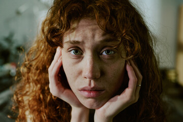 Portrait of a young woman with curly red hair resting her face in hands, gazing with somber...