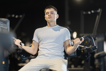 Concentrated young guy performing upper body workout on exercises machine in gym, doing bicep...