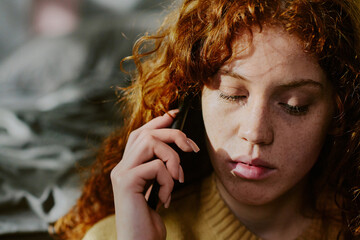 Red-haired woman listening intently while talking on smartphone indoors. Light casting soft shadows on her face, showcasing deep concentration in a cozy environment