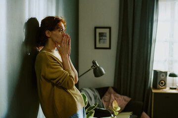 Woman standing by wall in living room, crying. Room decorated with lamp, framed photo, and few...