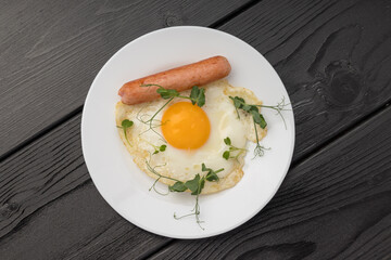 A fried egg with a sausage and green sprouts on a white plate, dark backdrop.