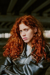 Portrait of red-haired woman standing under bridge, wearing leather jacket with intense gaze and...