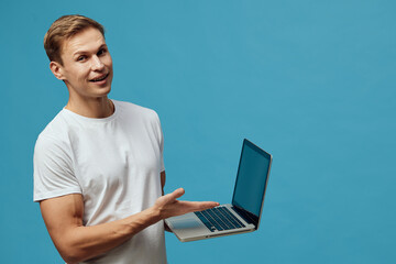 Smiling young man holding laptop pointing screen looking at camera casual white t-shirt isolated blue background digital nomad concept