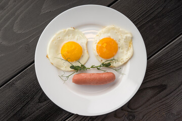 Fried eggs garnished with microgreens beside a sausage on a white plate, dark wooden background.