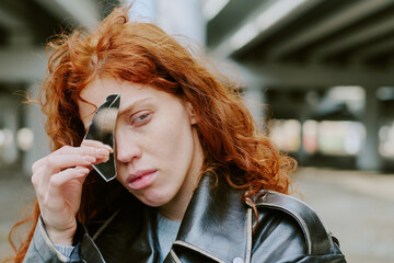 Woman with long red hair holding broken mirror fragment to face under bridge. Expressing intense...