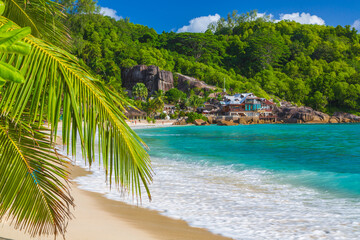 Sandy beach with palms and blue water of the Indian Ocean and volcanic stones with a hotel in the background. Island Mahe, the Seychelles