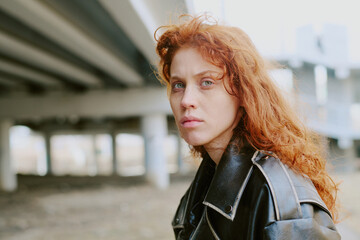 Portrait of woman with striking curly red hair, wearing black leather jacket and standing under...