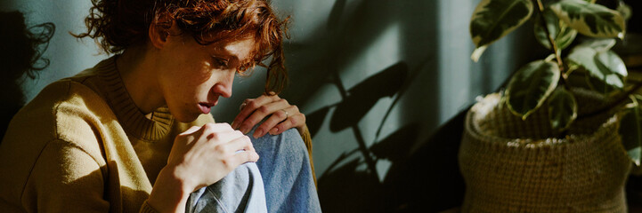 Person with red hair sitting by plant, contemplating something in sunlight. Sunlight casting...