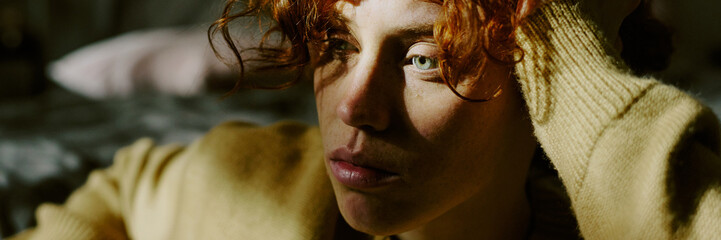 Young woman with curly red hair gazing thoughtfully, with sunlight highlighting her features. Warm...