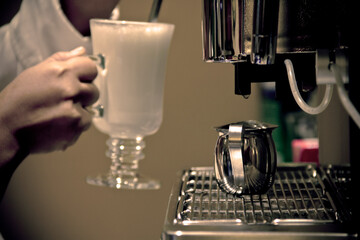 Close-up of a Hispanic barista’s hand holding a classic glass cappuccino cup near a polished...