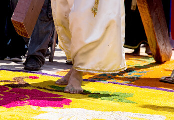 Barefoot figure of Jesus carrying the cross walks over a vibrant sawdust carpet during a...