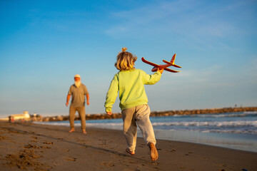 Little boy runs barefoot on beach holding orange toy airplane with grandfather watching in background, playful moment at seaside.