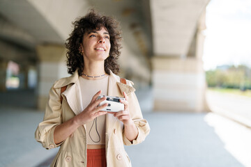 Smiling Mediterranean Female Tourist Exploring Urban Sights Holding Camera Under a Bright Sunlit...