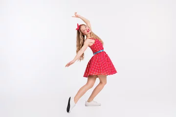 Gordijnen Young woman in red polka dot dress dancing against white background, evoking retro charm and lively energy © deagreez