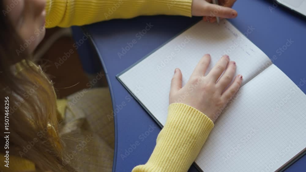 Poster A person is writing in a notebook with graph paper pages. They are wearing a yellow sweater and writing on a blue desk. The focus is on the hand holding the pen and the open notebook.