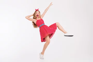 Selbstklebende Fototapeten Joyful young woman in a nostalgic red polka dot dress dancing energetically against a white backdrop, celebrating retro style © deagreez